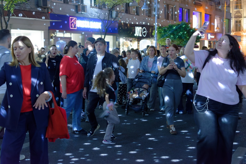 10 - A pedestrian crowd gathered on a city street at night, illuminated by floating or projected light spots, with the storefront signs of Vista Bank and the clothing store JOYZ